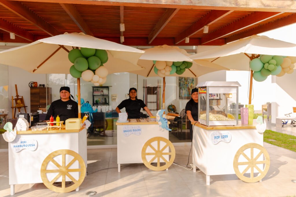 Tres carritos de comida temáticos para bodas (Mini Hamburguesas, Snacks Dulces, Palomitas) con paraguas y decoración de globos en una terraza moderna.