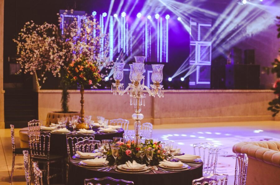 Decoración de mesas para eventos de gala en algarabiafiestas.com, con candelabros de cristal, centros de mesa florales y mobiliario transparente bajo iluminación neón profesional.